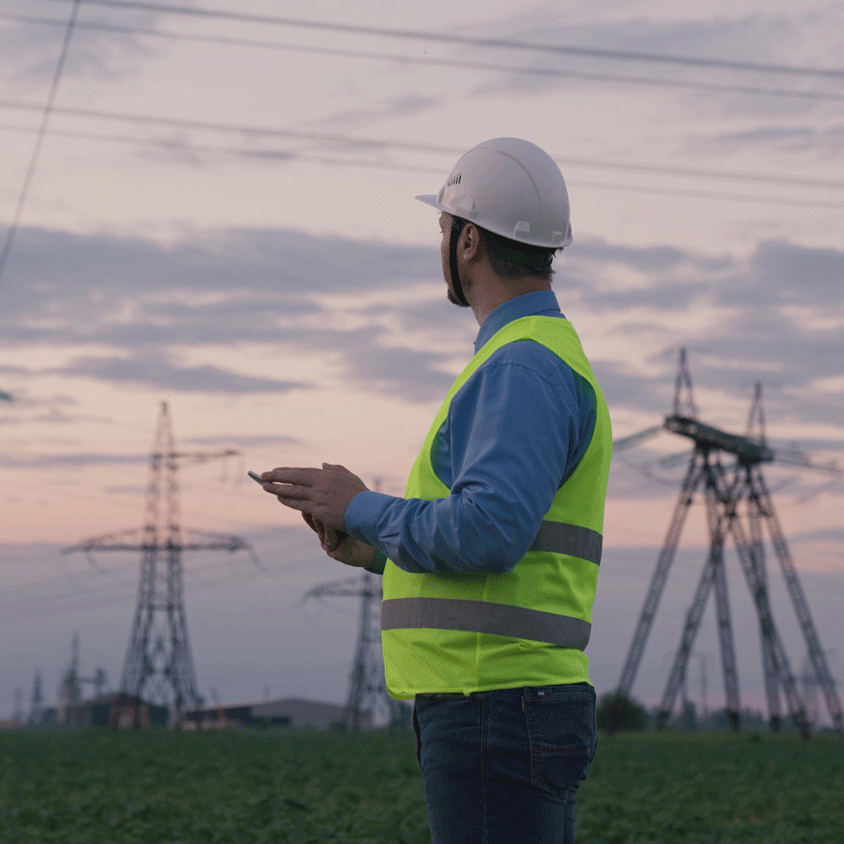 Stromnetztrennung Mann in Sicherheitsweste und Helm steht vor Strommasten bei Sonnenuntergang.